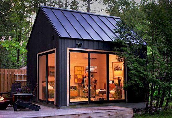 modern black detached backyard office with a metal roof, large glass doors, and warm interior lighting